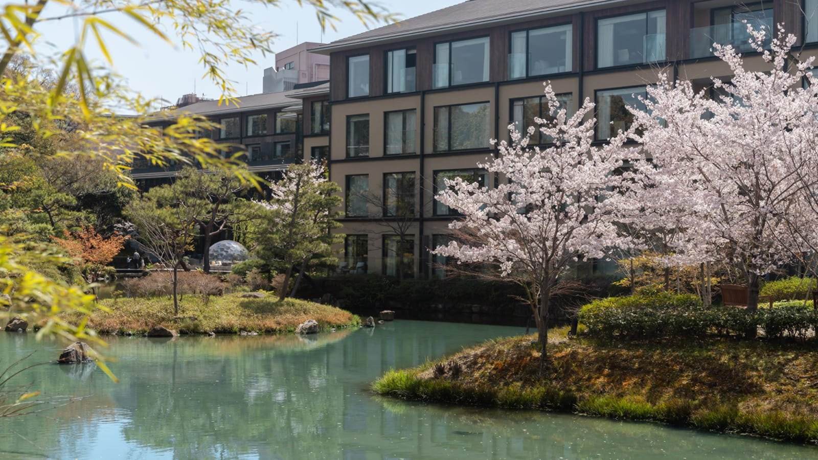 Etang japonais bordé d'érables et de pierres, avec un pavillon de thé en bois clair et la façade contemporaine d'un hôtel à l'arrière-plan