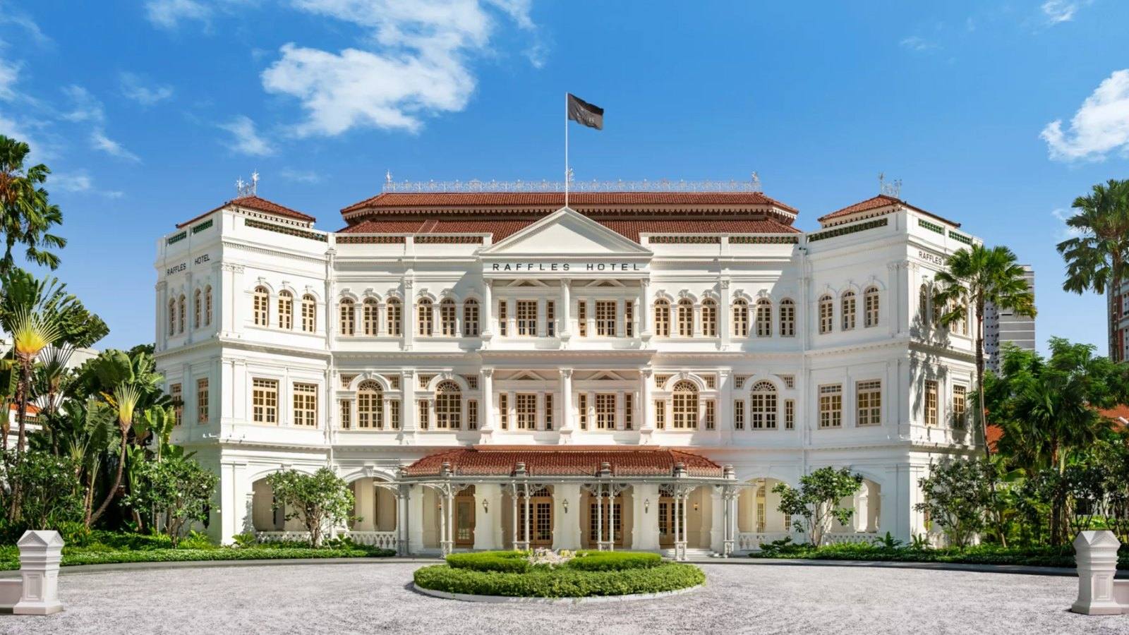 Façade blanche à arcades et vérandas du Raffles Singapour vue depuis Beach Road, avec palmiers en premier plan et tours de bureaux en arrière-plan.