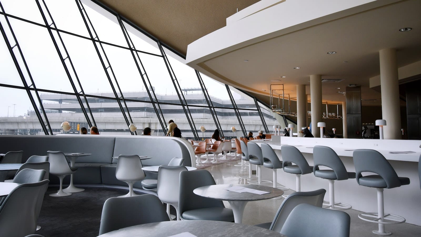 Salle du Senator Lounge de Francfort : rangées de fauteuils gris devant une baie vitrée donnant sur le tarmac, tables basses, passagers travaillant sur ordinateur portable.