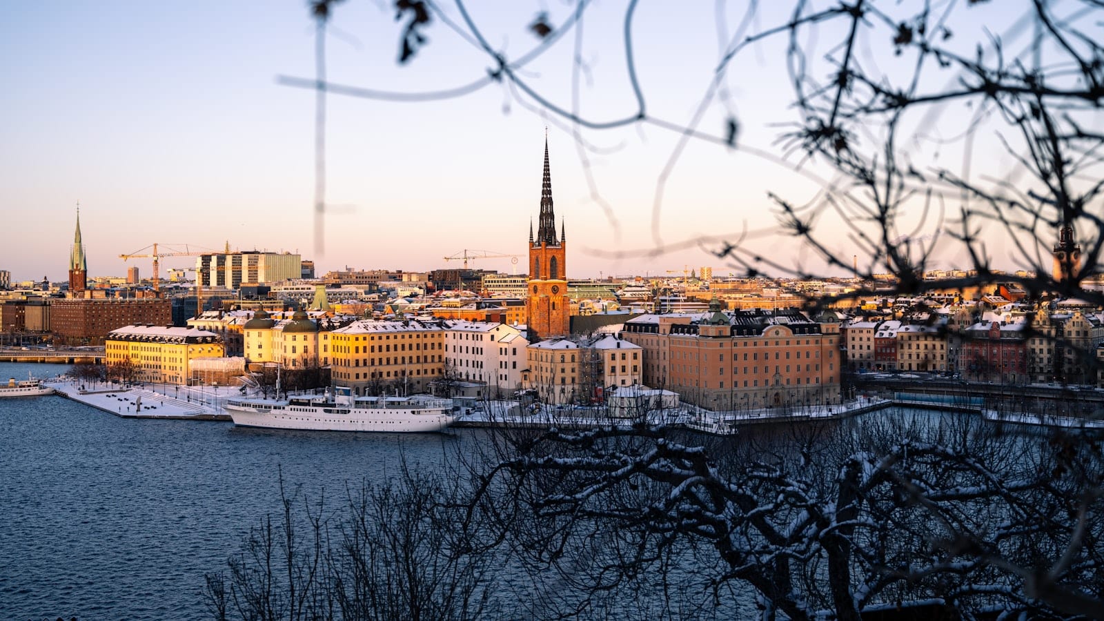 Une rue de Gamla Stan à Stockholm en fin d’après-midi d’hiver, façades ocre éclairées par des lampes de vitrine, neige tassée sur les pavés et ciel déjà bleu nuit.